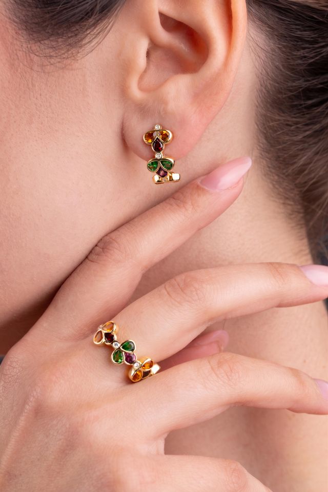 A close-up lifestyle shot of a model wearing a matching pair of gold earrings and ring set with green, red, and white gemstones details and yellow gold.