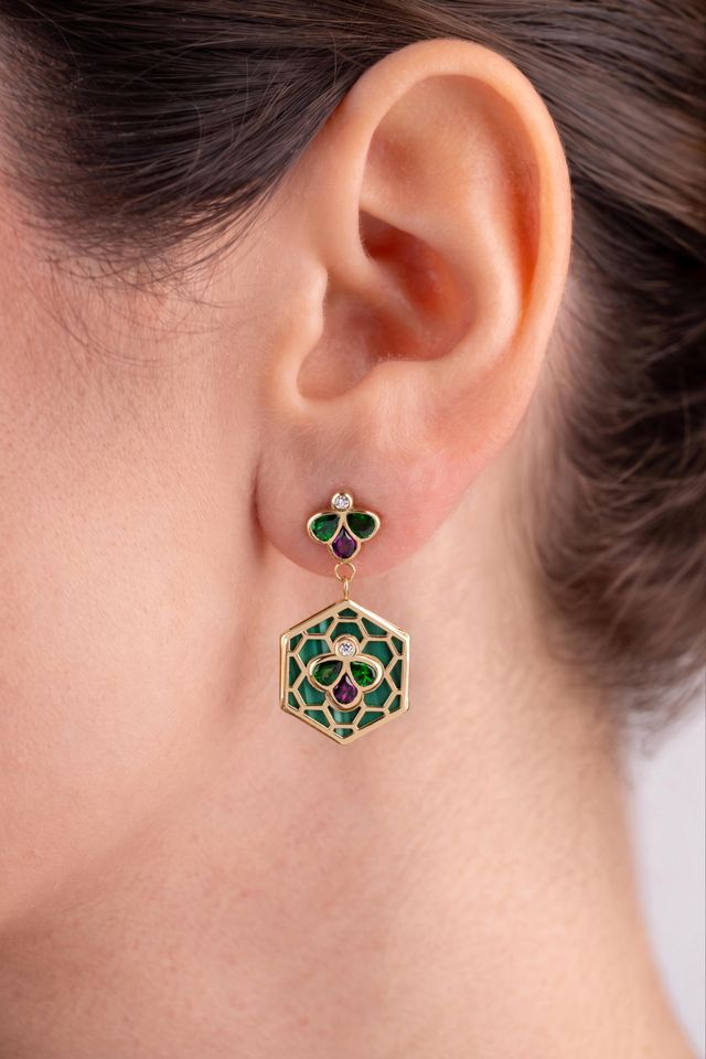 A close-up of a model's ear wearing a gold dangle earring with green and purple enamel details and a geometric hexagonal drop.