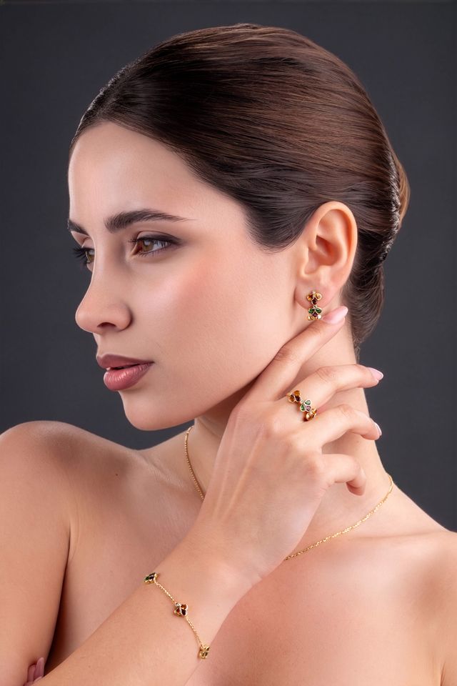 A woman with dark hair styled in an updo models a matching gold jewelry set, including a delicate chain necklace, bracelet, and earrings with small green gemstone accents, against a dark gray background.