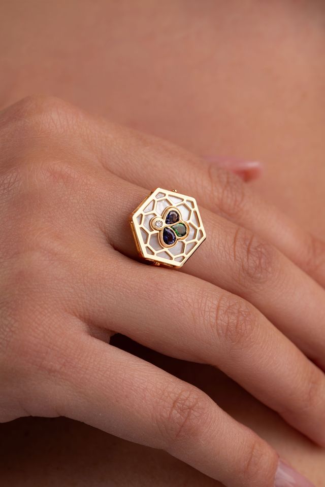 A gold hexagonal ring with a floral openwork design and white enamel details, shown worn on a person's finger.