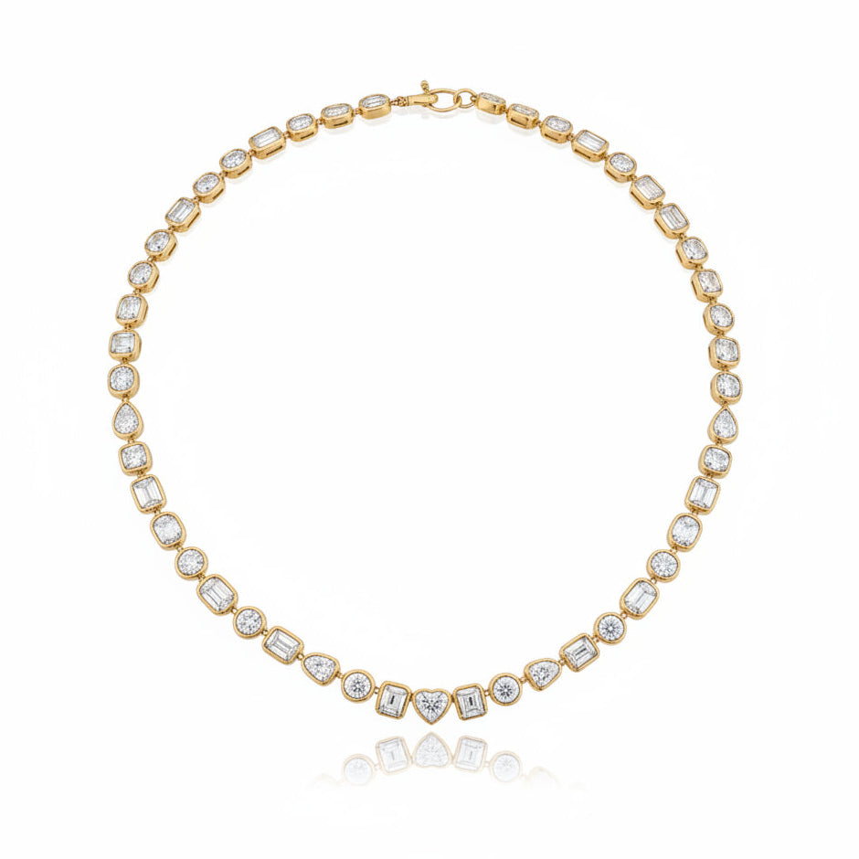A yellow gold tennis necklace featuring multi-shaped diamonds in bezel settings, arranged in a circular shape on a white background with a soft reflection below.