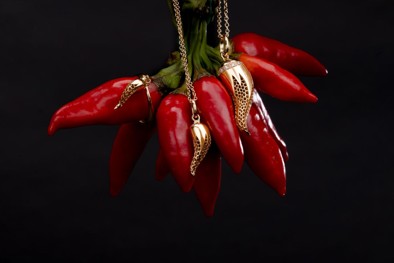 Red chili peppers hanging from a chain with gold-toned pendant charms shaped like stylized chili peppers, set against a solid black background.
