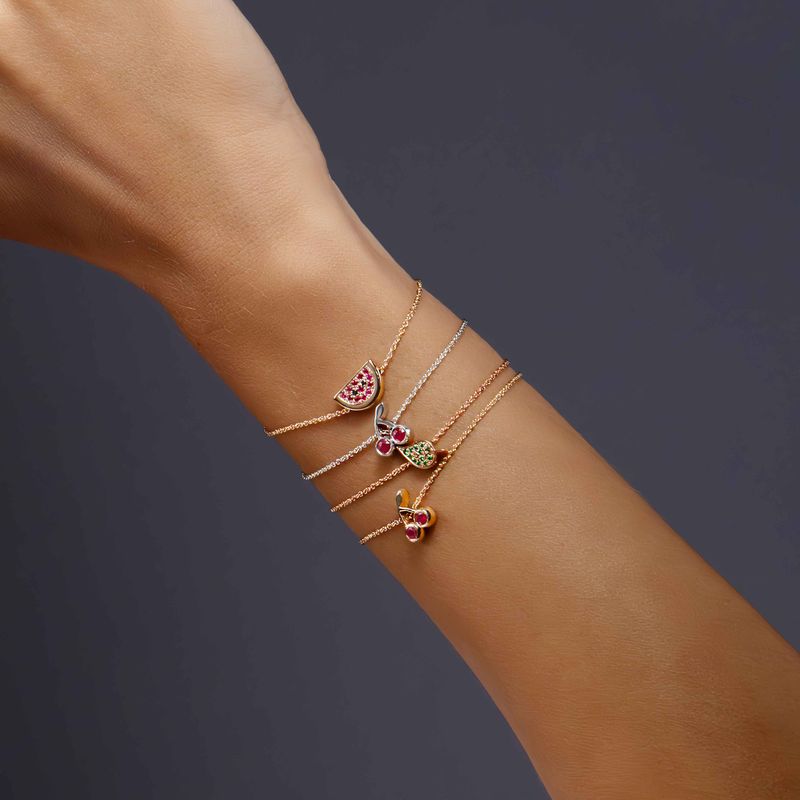 A close-up of a person's wrist wearing three delicate rose and white gold bracelets stacked together. Each bracelet features a small colorful  charm, including pink, green, and yellow details, set against a dark gray background.