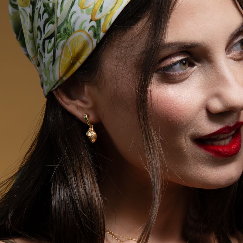 A close-up of a woman wearing a green and white lemon-patterned headscarf and a small gold dangle earring, set against a warm brown background.