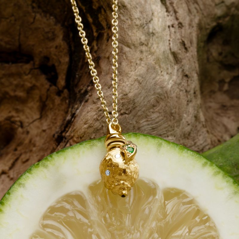A gold pendant necklace featuring a textured gold lemon shaped pendant with a small green gemstone and a tiny white diamonds, hanging in front of a sliced lime on a rustic wood background.