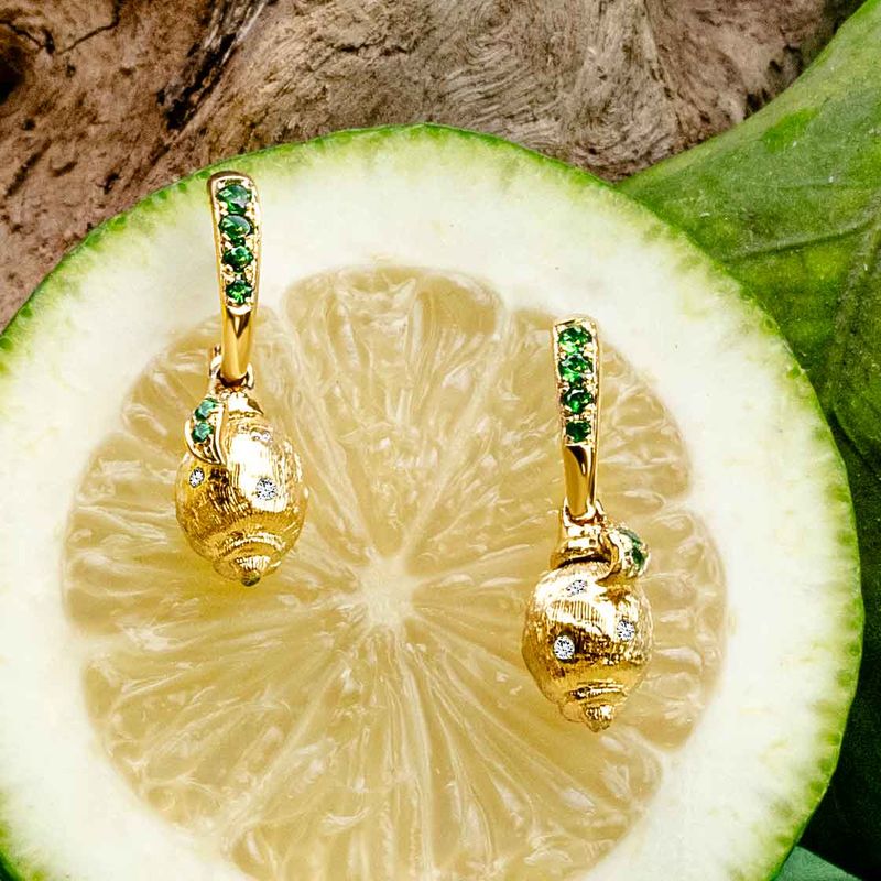 A pair of yellow gold drop earrings with green gemstone accents and white diamonds, displayed on a sliced lime over a rustic wood background.