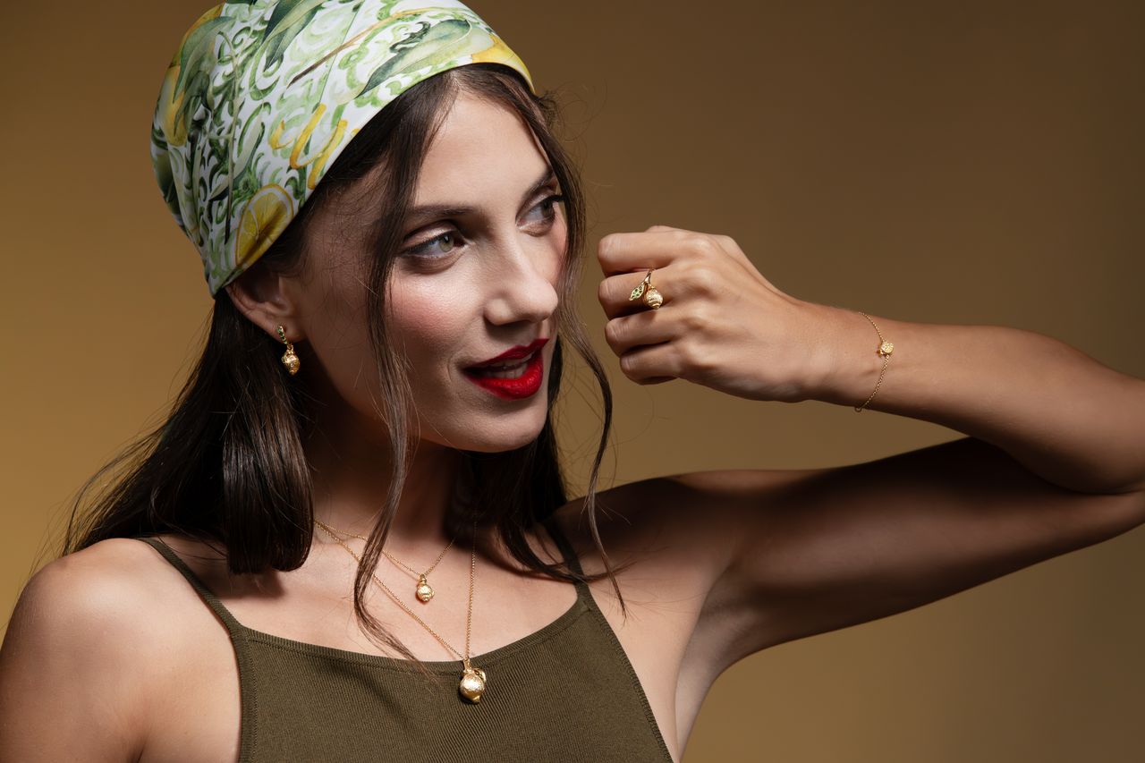 A woman wearing a green and yellow patterned headscarf, gold jewelry including a necklace, earrings, and ring, and a dark olive green top, posing against a warm brown studio background.