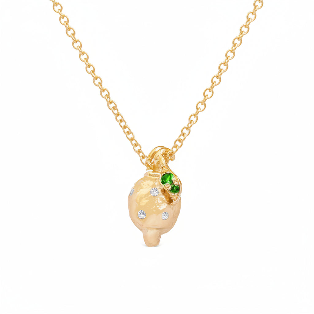 A gold pendant necklace featuring a stylized, textured lemon pendant with small white diamonds and a green emerald color accent gemstones, shown on a white background.