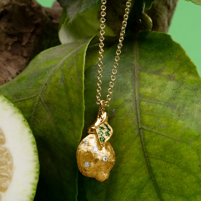 A gold pendant necklace shaped like a lemon, featuring green gemstone accents and tiny white diamonds, hanging from a gold chain against a background of green leaves and a sliced lemon.