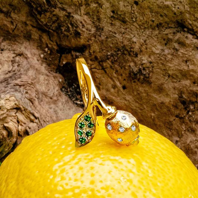 A gold ring designed as a stylized citrus fruit with a leaf and a gemstone-covered sphere, resting on a bright yellow lemon against a textured brown rock background.