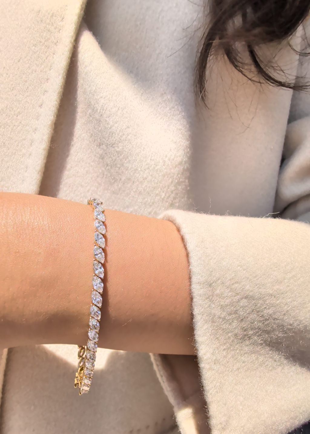 A close-up of a person's wrist wearing a gold tennis bracelet with marquise diamonds, styled with a cream-colored coat.