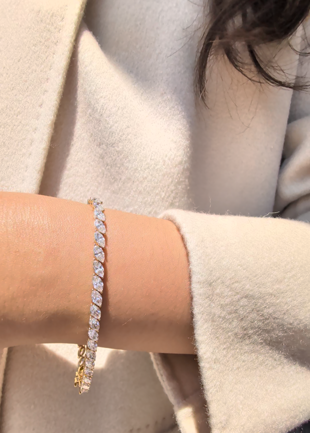 A close-up of a person's wrist wearing a gold tennis bracelet with marquise diamonds, styled with a cream-colored coat.