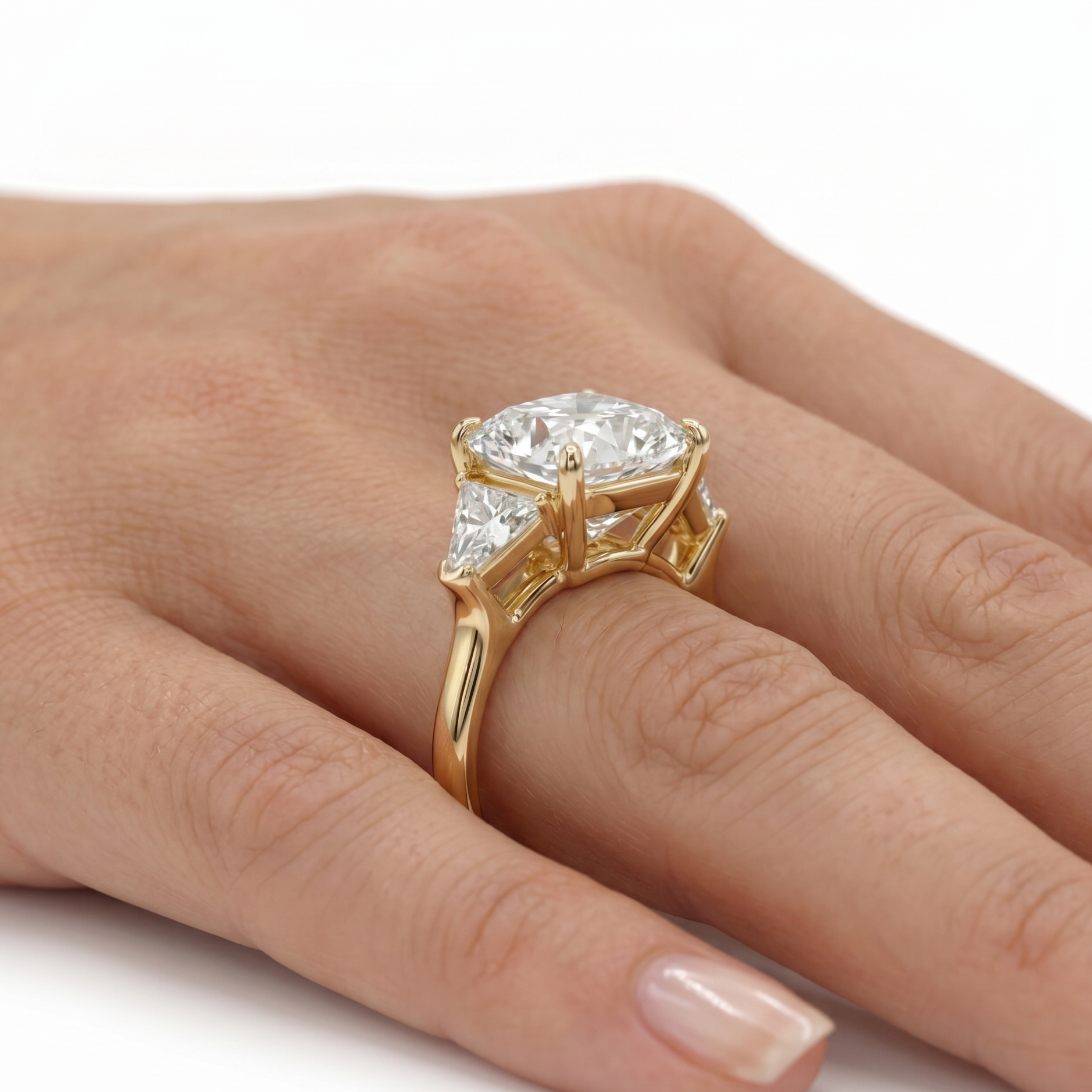 A yellow gold three-stone ring featuring a large Cushion brilliant-cut center diamond flanked by two smaller trillion lab grown diamonds, set against a plain white background an models hand.

