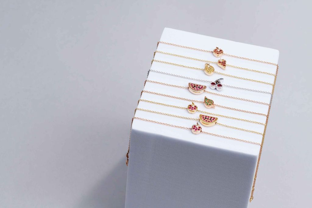A stack of delicate gold-tone bracelets with small fruit charms, displayed on a white rectangular block against a light gray background.


