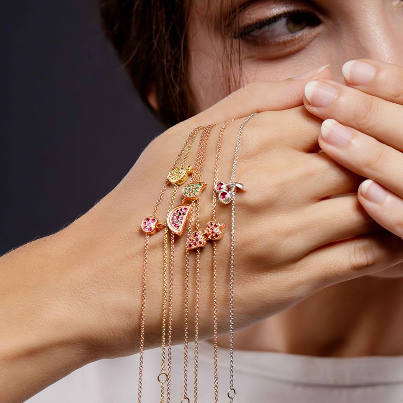 A close-up lifestyle shot of a person holding their hand near their face, showcasing several delicate rose and white and yellow gold chain bracelets with colorful enamel flower charms.