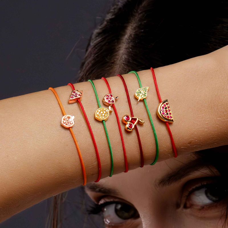 A close-up of a person's forearm wearing six thin charm bracelets in orange, red, green, and gold, each with a small decorative charm including a strawberry, lemon, leaf,apple, mango, and watermelon motif.