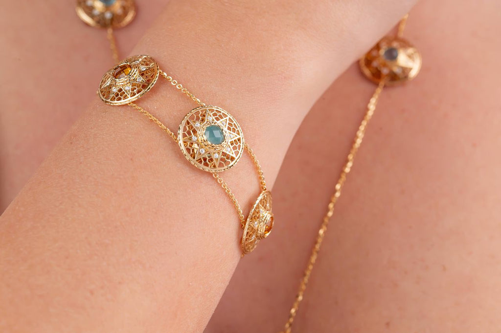 A gold chain bracelet featuring four ornate circular filigree medallions, each set with a light blue or yellow stone and a central gold bead shown worn on a persons wrist.