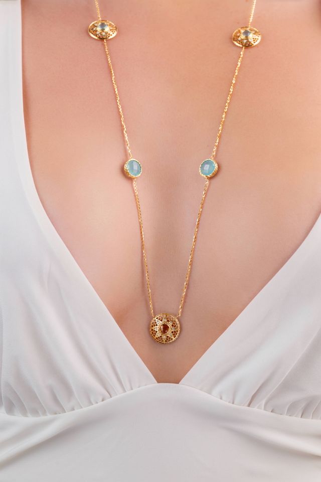A gold chain necklace with circular light blue and red gemstone accents, shown worn on a model's neck over a white V-neck top.