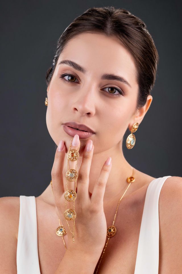 A woman modeling a gold jewelry set featuring a delicate chain with round clear stones draped across her hand, matching drop earrings, and a necklace, set against a dark gray background.