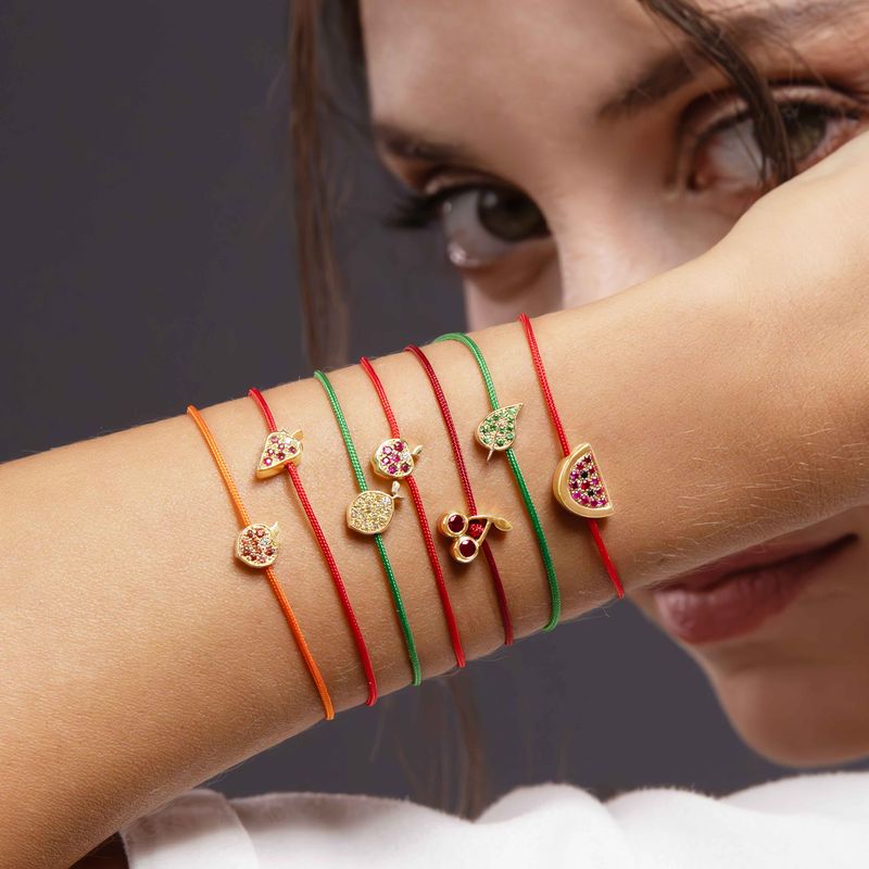 A close-up lifestyle shot of a woman's wrist wearing six colorful string bracelets with gold-toned leaf and fruit charms, including a cherry, watermelon slice, lemon and leaf designs.