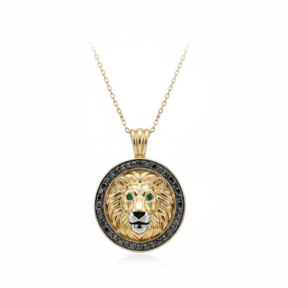 A round lion head pendant necklace in yellow gold with a halo of small black diamonds around the border and green gemstone eyes, suspended from a delicate gold chain on a white background.