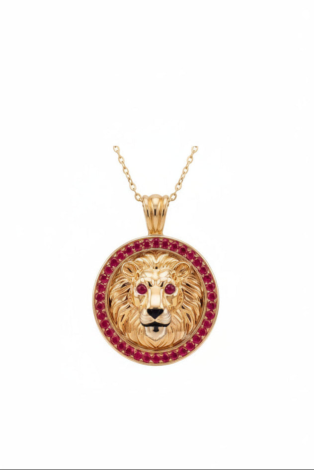 A gold lion head pendant necklace on a delicate gold chain, featuring a border of red ruby gemstones around the lion's mane and ruby-red eyes, set against a plain white background.