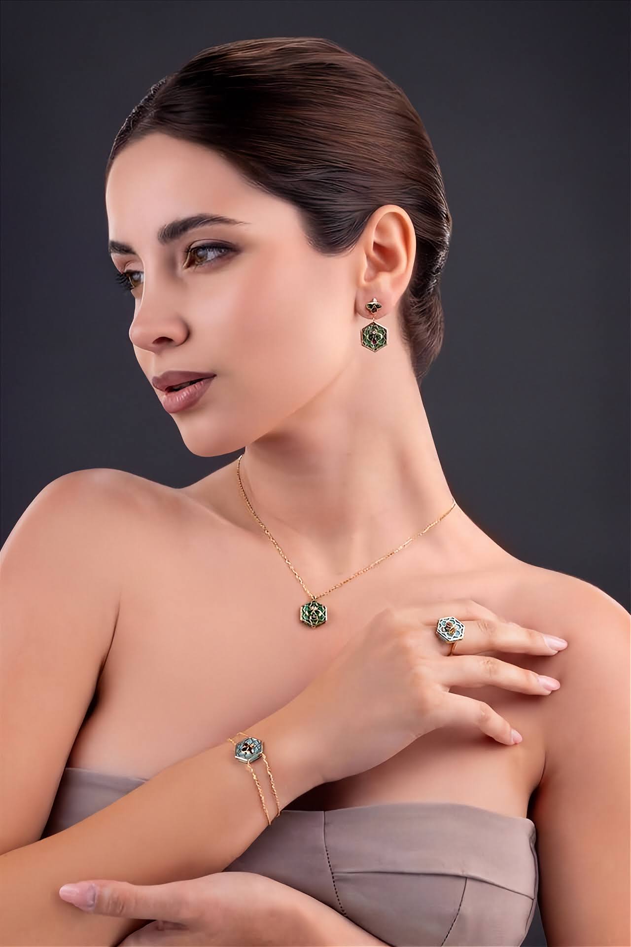 A woman models a coordinated jewelry set featuring a gold necklace, matching bracelet, and ring with green gemstone accents, against a dark gray studio background.
