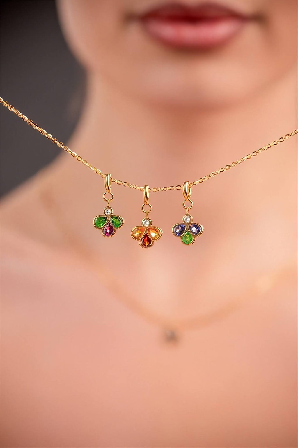 A close-up lifestyle shot of a model showing a delicate gold chain necklace with three small, colorful flower-shaped charms (green, red, orange, and blue) set against a soft-focus background of the model's face and neck.