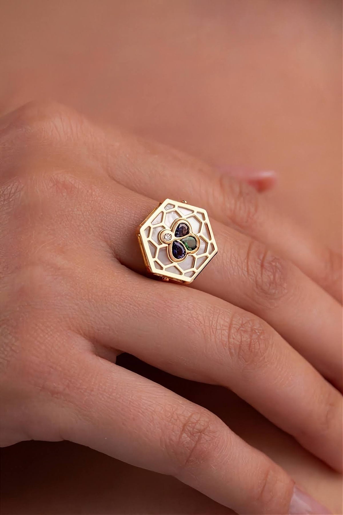 A gold hexagonal ring with a floral openwork design and white enamel details, shown worn on a person's finger.