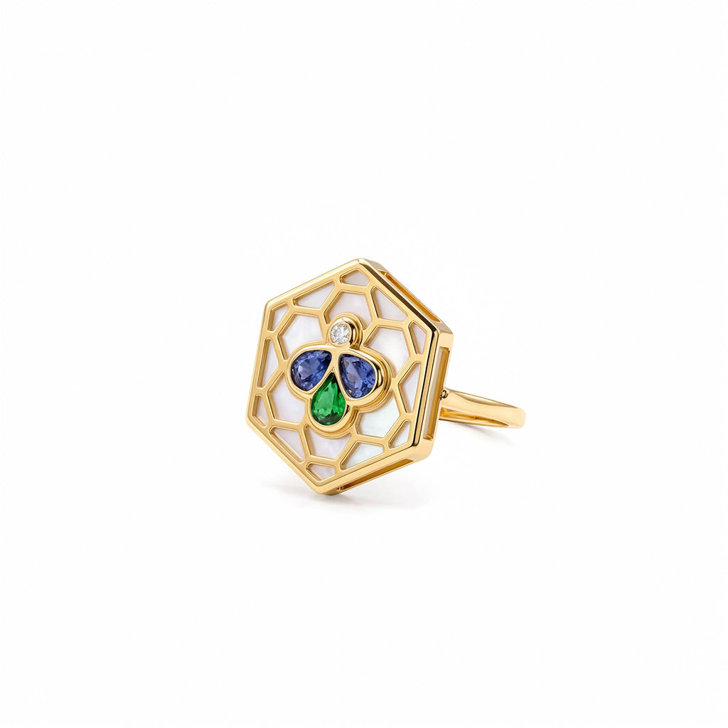A gold hexagonal ring featuring an openwork honeycomb pattern with white enamel inlay and a central clover motif made of blue and green stones, accented by a small clear gemstone.
