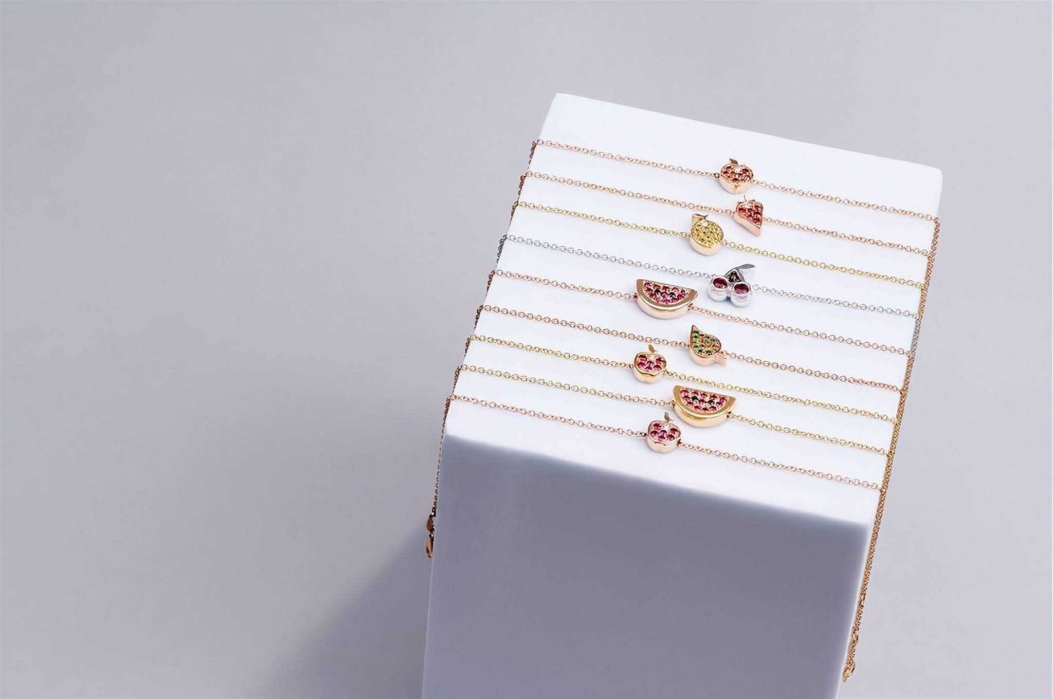 A stack of delicate rose and yellow 
gold with small fruit-themed charms, displayed on a white rectangular block against a light gray background.