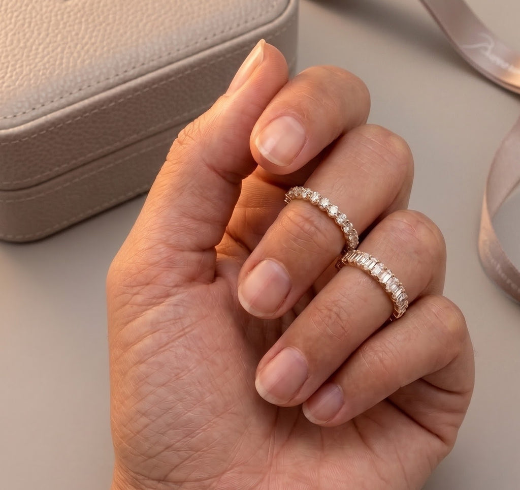A close-up of a hand wearing two thin diamond eternity rings  in yellow gold with a textured beige jewelry box in the background.