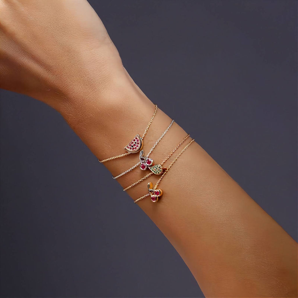 A close-up of a person's wrist wearing three delicate rose and white gold bracelets stacked together. Each bracelet features a small colorful  charm, including pink, green, and yellow details, set against a dark gray background.