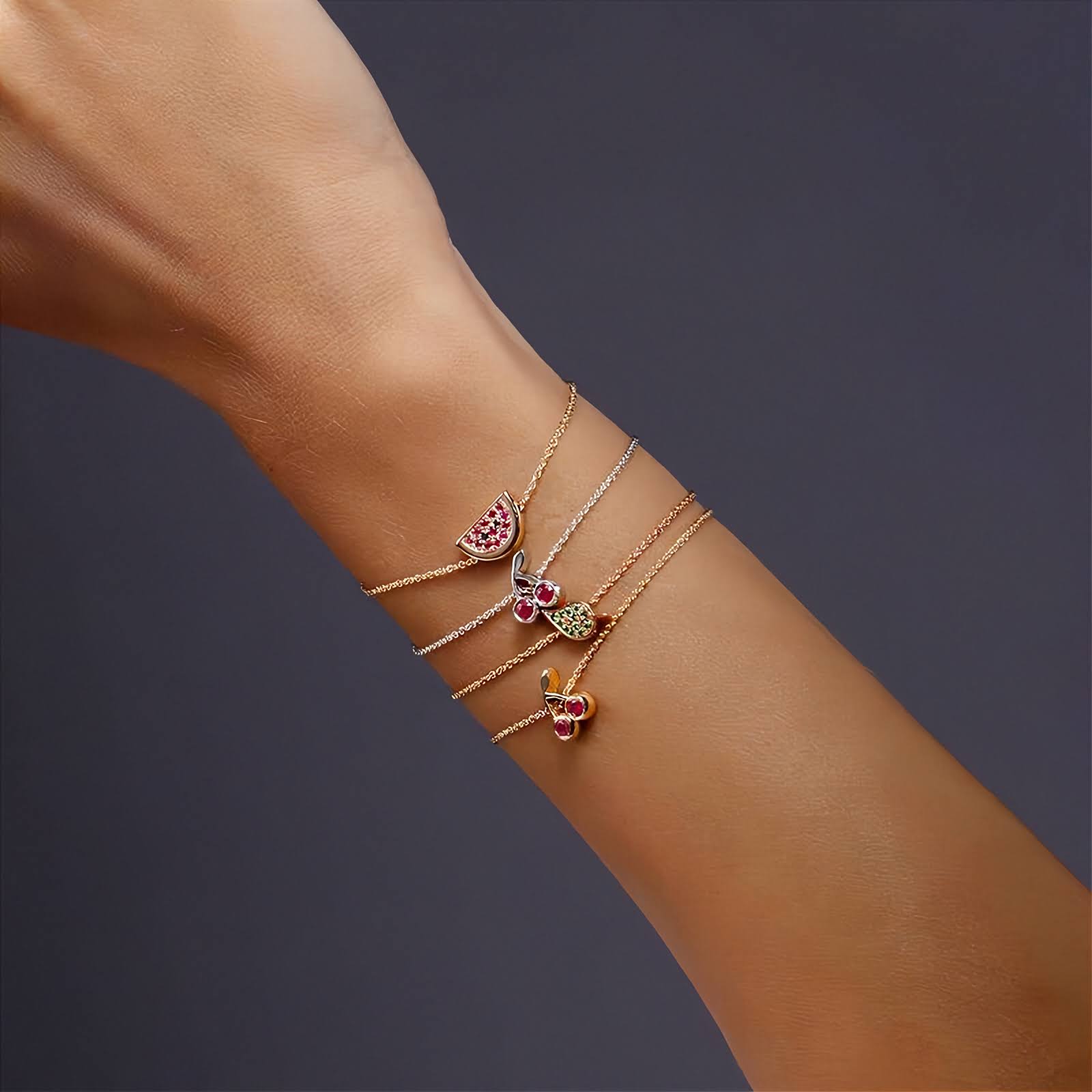 A close-up of a person's wrist wearing three delicate rose and white gold bracelets stacked together. Each bracelet features a small colorful  charm, including pink, green, and yellow details, set against a dark gray background.
