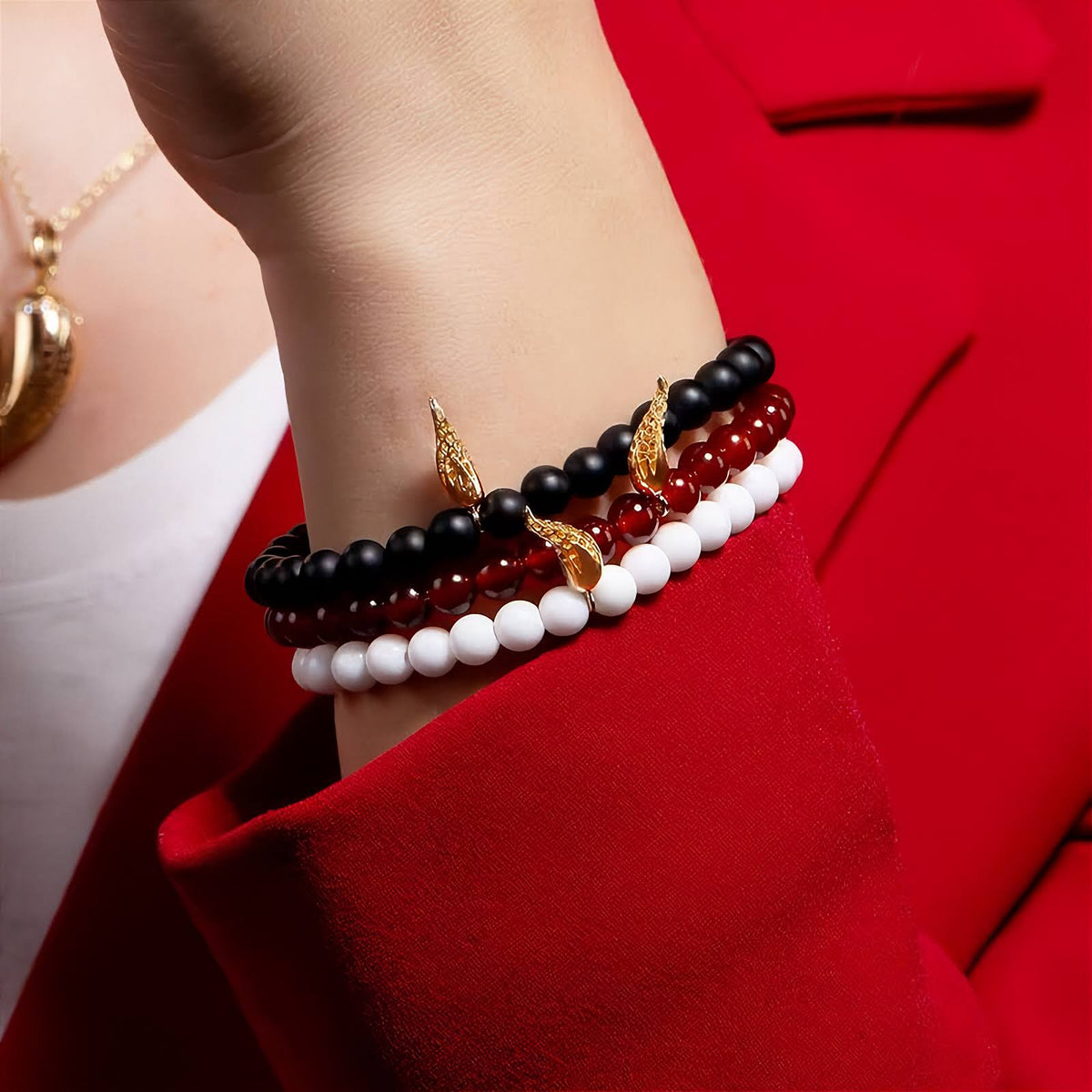 Corno Charm Bracelet with 18k Yellow Gold Rosmundo Italian Corno Charm Bracelet with Onyx, Carnelian Beads and white agate beads. A close-up of a person's wrist wearing three beaded bracelets: one black, one white, and one translucent dark red, each accented with a small gold horn charm. The person is wearing a bright red blazer and white top.
