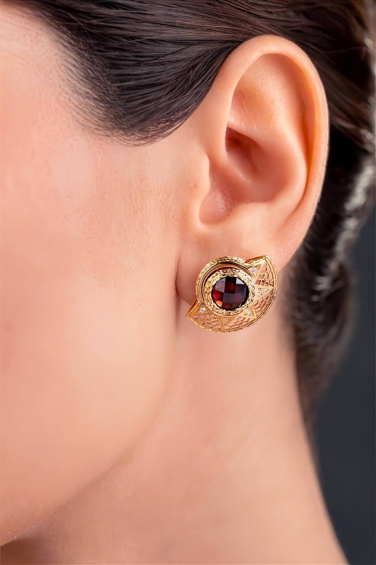 A pair of gold filigree stud earrings featuring round deep red garnet centers and small clear accent stones, shown on a model.