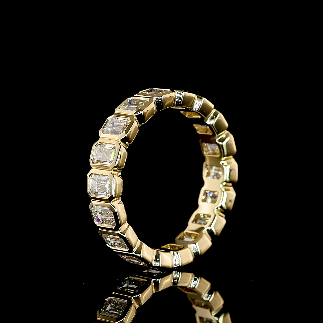 14K yellow gold bezel set emerald cut lab grown diamond eternity band ring 2.67 ctw Inga Fine Jewelry shown from the side on black background.