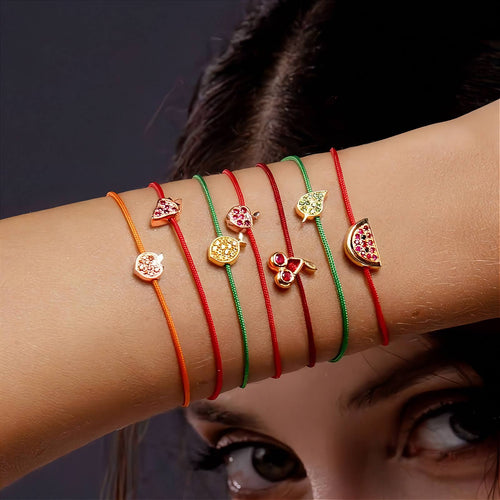 A close-up of a person's forearm wearing six thin charm bracelets in orange, red, green, and gold, each with a small decorative charm including a strawberry, lemon, leaf,apple, mango, and watermelon motif.