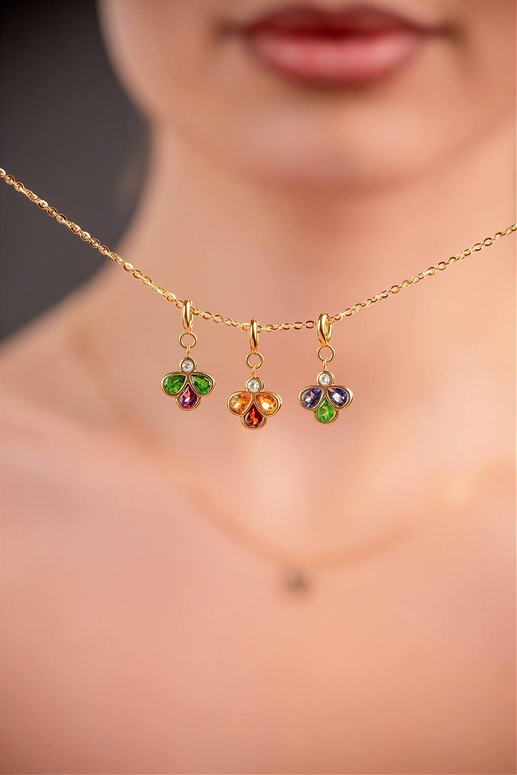 A close-up lifestyle shot of a model showing a delicate gold chain necklace with three small, colorful flower-shaped charms (green, red, orange, and blue) set against a soft-focus background of the model's face and neck.