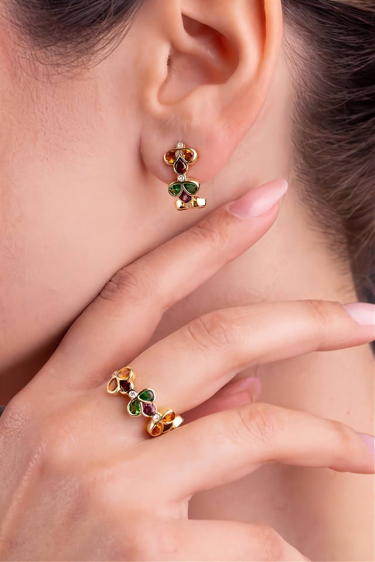 A close-up lifestyle shot of a model wearing a matching pair of gold earrings and ring set with green, red, and white gemstones details and yellow gold.