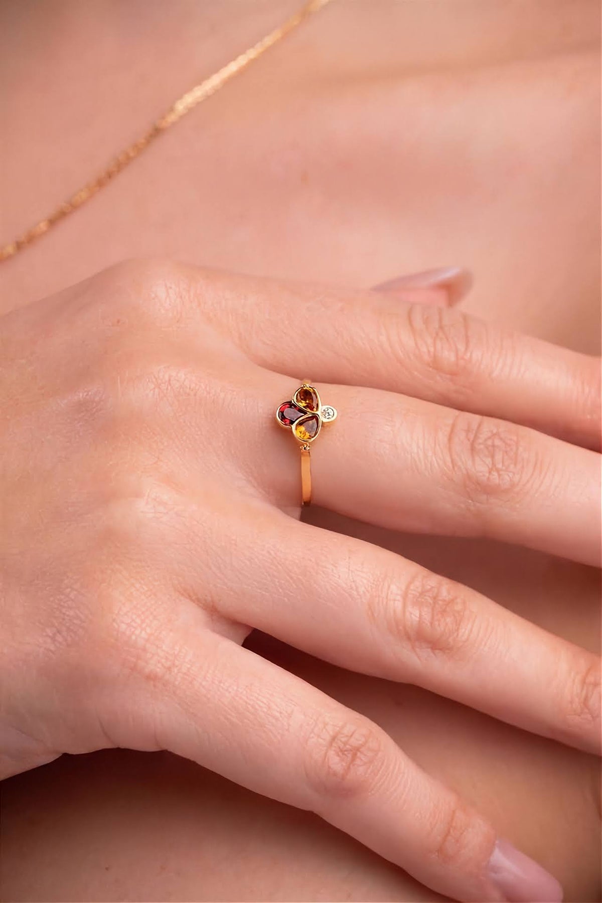 A yellow gold ring featuring three gemstone clusters: a top round white diamond, a middle pear-cut two orange stone, and a bottom teardrop-cut dark red stone, set on a polished gold band shown on a model.