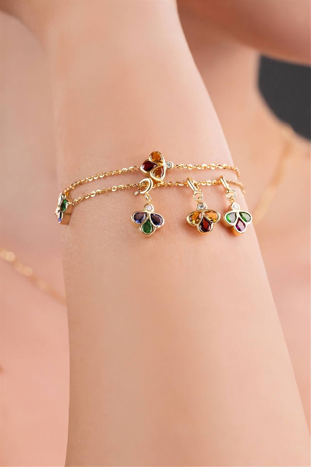 A close-up of a person's wrist wearing two delicate gold bracelets adorned with colorful charms.