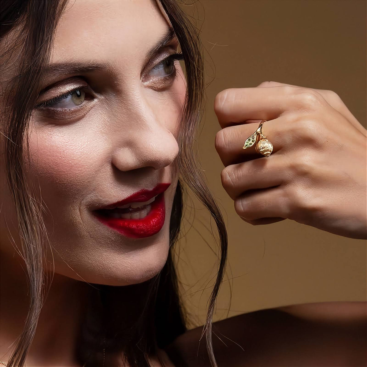 A gold ring featuring a textured lemon design and a leaf-shaped top set with green stones and small round diamonds, shown on a plifestyle image on the models finger.