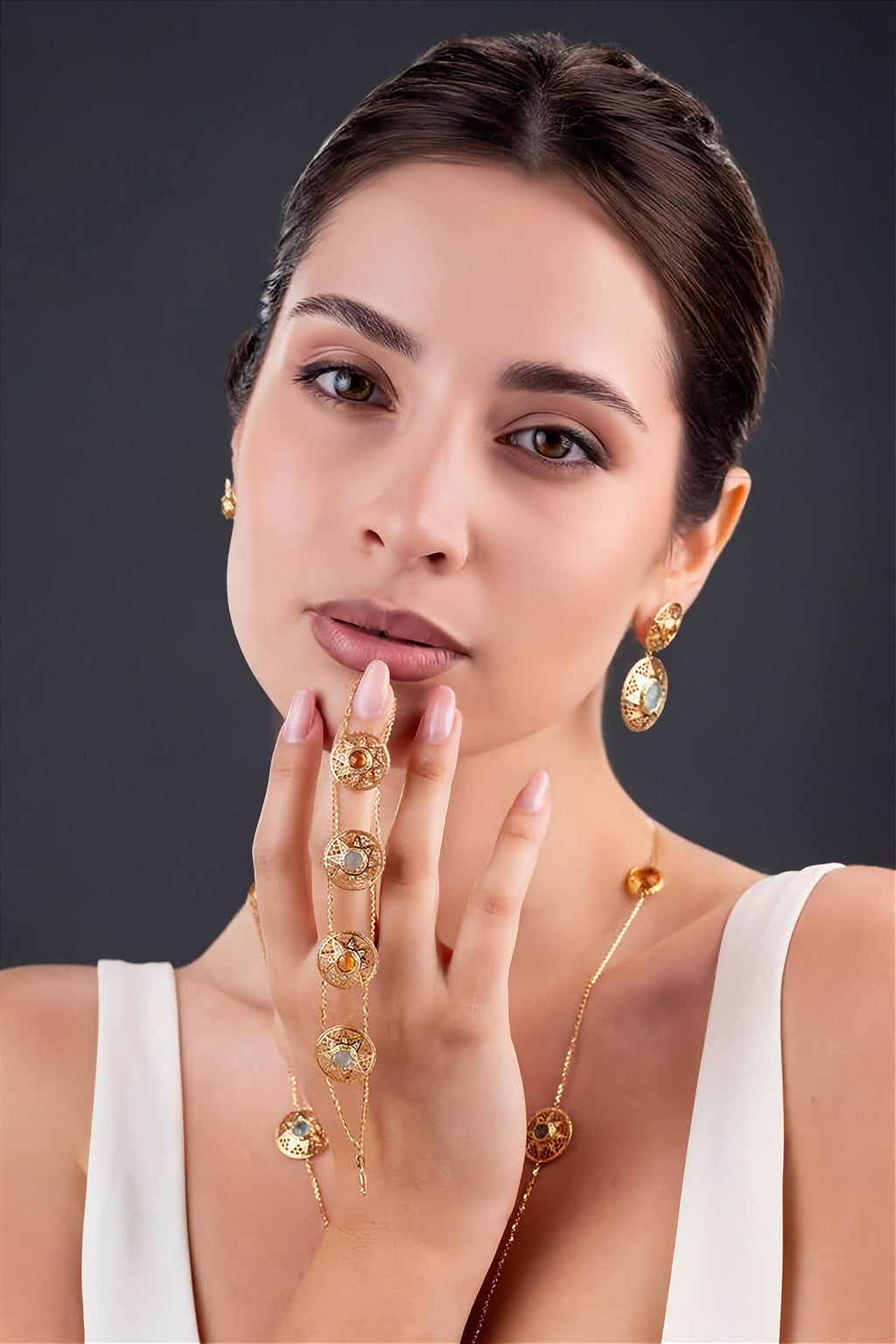 A woman modeling a gold jewelry set featuring a delicate chain with round clear stones draped across her hand, matching drop earrings, and a necklace, set against a dark gray background.