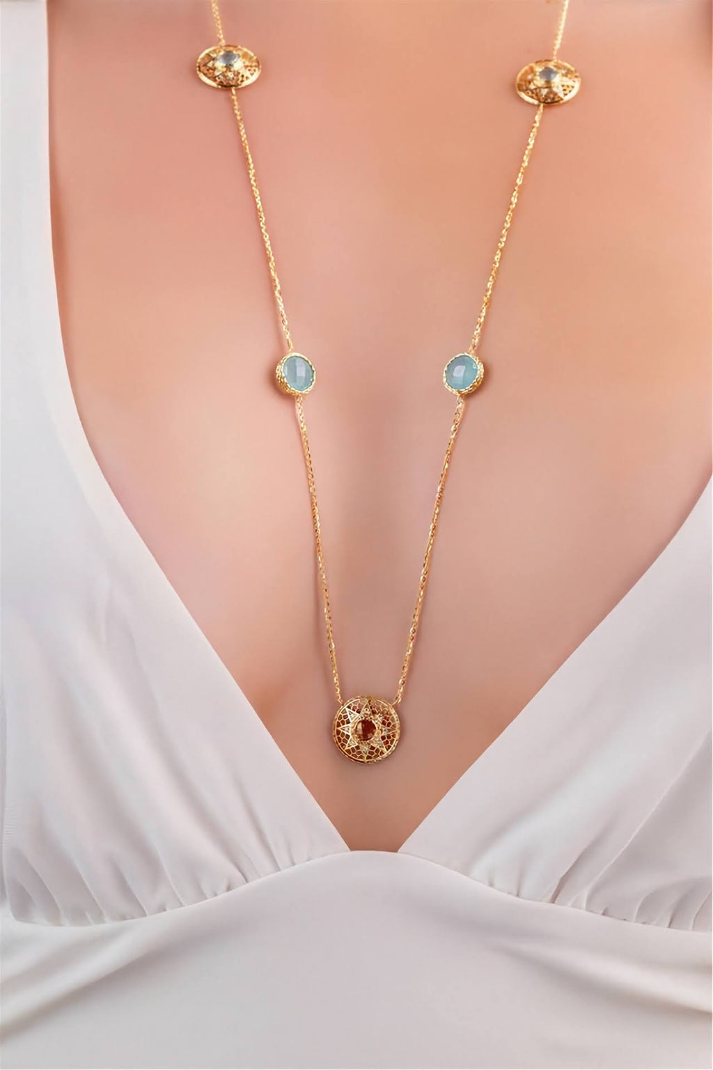 A gold chain necklace with circular light blue and red gemstone accents, shown worn on a model's neck over a white V-neck top.