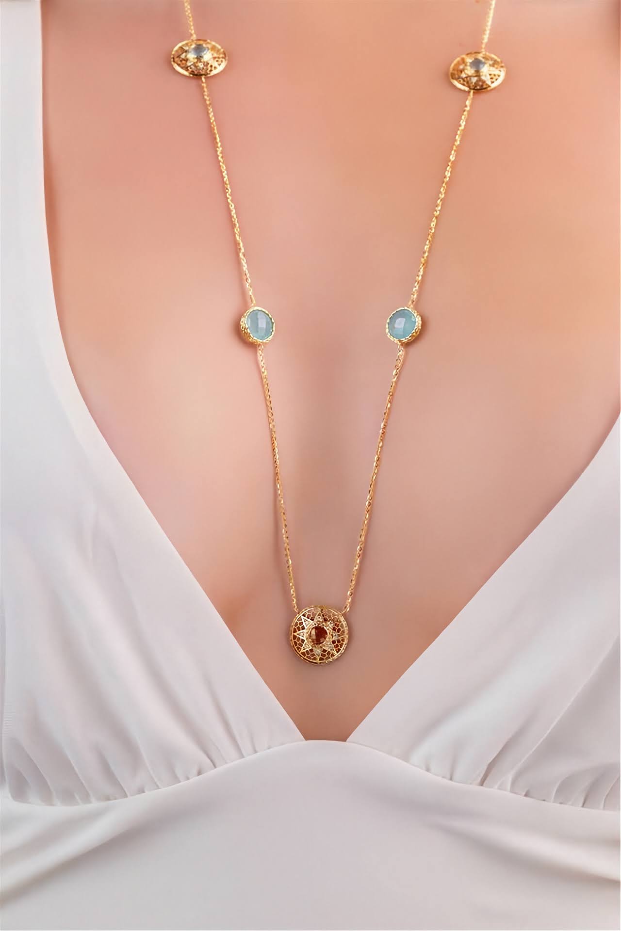 A gold chain necklace with circular light blue and red gemstone accents, shown worn on a model's neck over a white V-neck top.