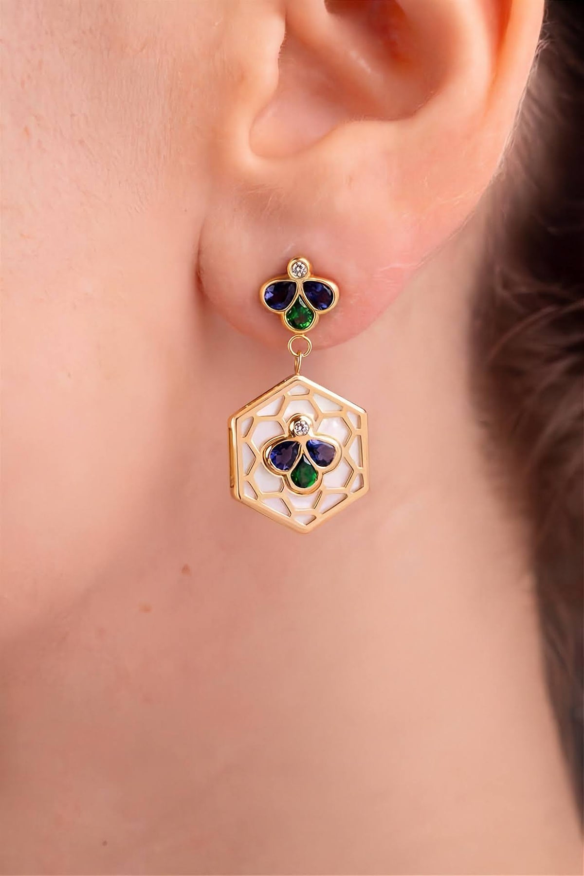 A close-up of a person's ear wearing a gold dangle earring with a hexagonal filigree drop and a small clover-shaped top set with green and dark blue stones.