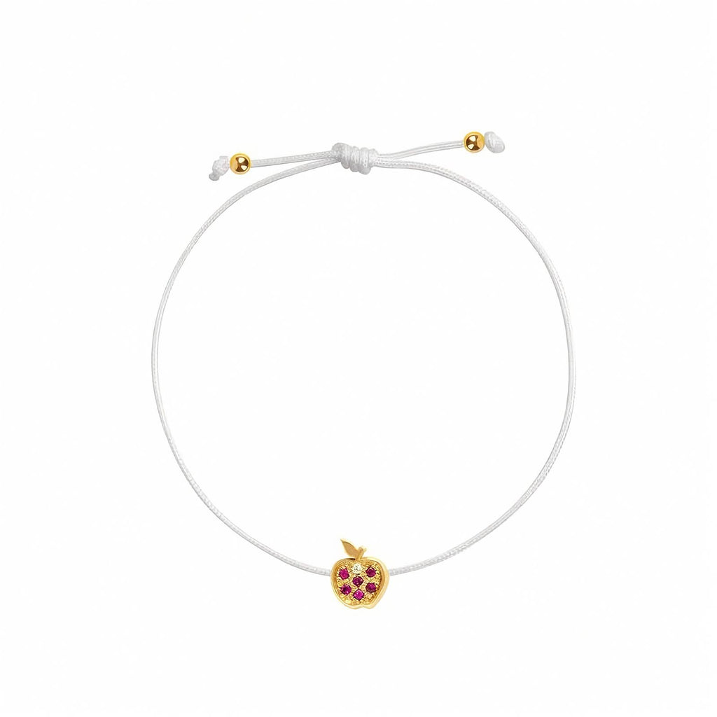 Rosmundo collaboration 18k yellow gold apple string bracelet with natural rubies and diamond on adjustable macrame cord. A white adjustable cord bracelet featuring a small yellow gold apple charm embellished with purple gemstones, set against a plain white background.