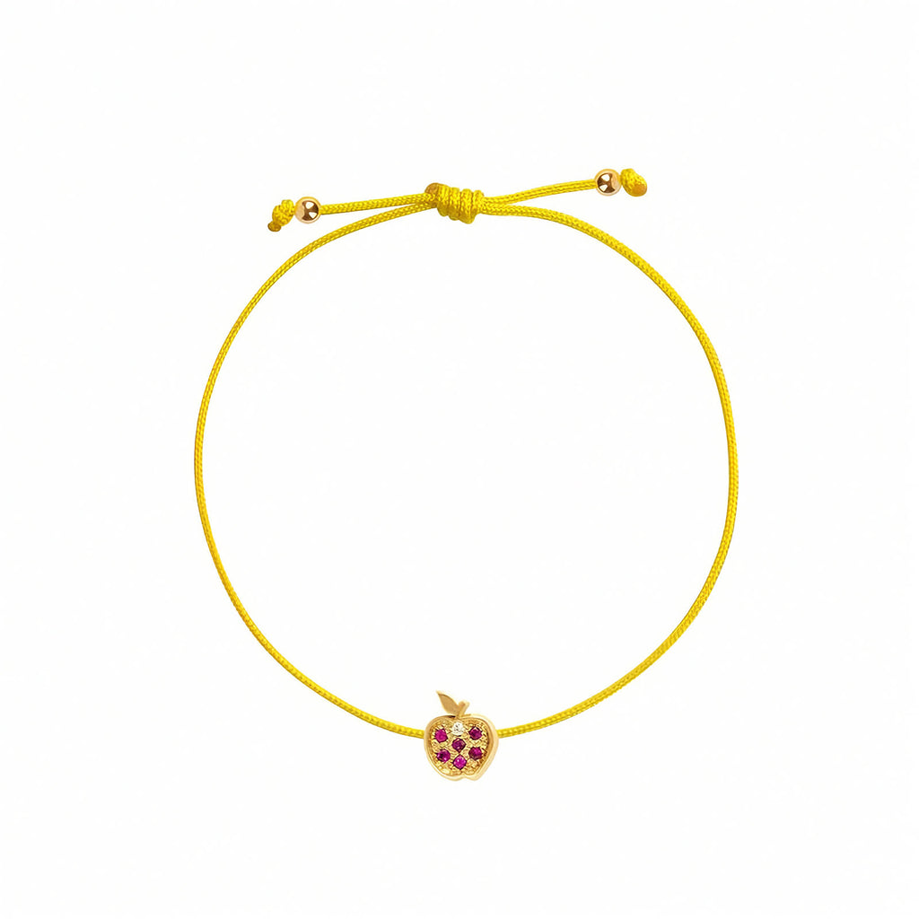 Rosmundo collaboration 18k yellow gold apple string bracelet with natural rubies and diamond on adjustable macrame cord. A green adjustable cord bracelet featuring a small yellow gold apple charm embellished with purple gemstones, set against a plain white background.